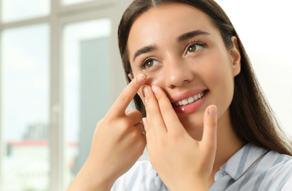 A young woman is smiling while she is putting in her soft contact lens at the tip of her finger in her right eye.