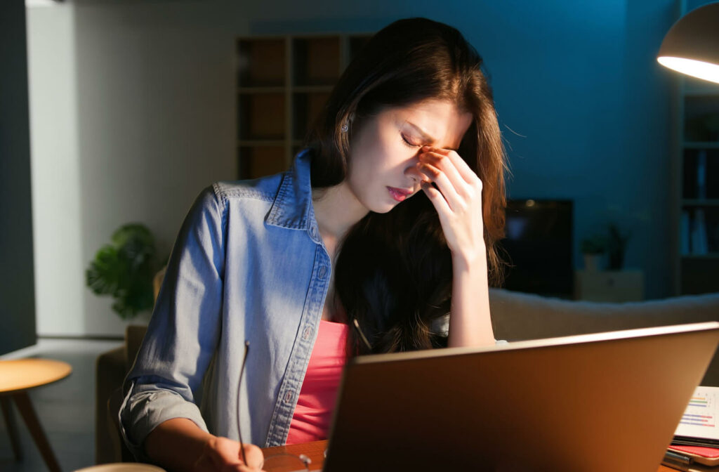 A young woman sitting in front of her computer in low light is massaging her eyes due to difficulty in seeing.