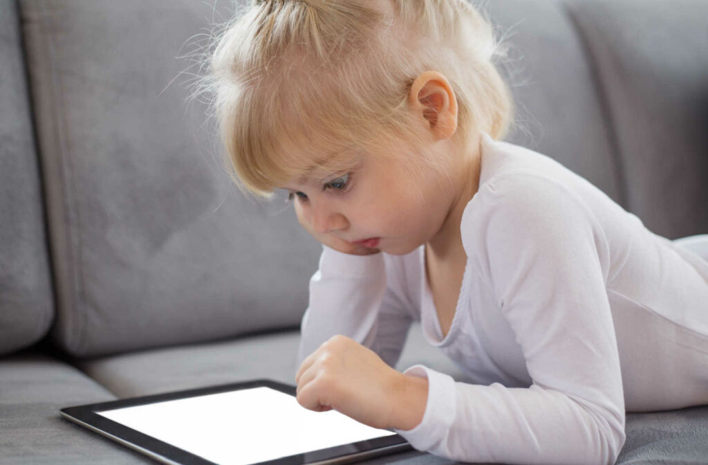 A female toddler laying on a couch playing on an electronic tablet.