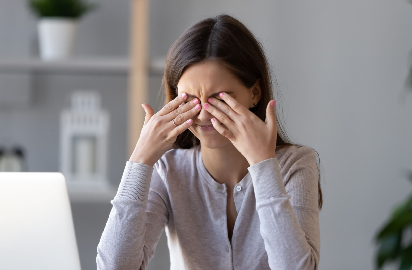 A woman with long, straight, brown hair wearing a gray button-up sweater is sitting in an office with a laptop off to the side, rubbing her eyes with both hands