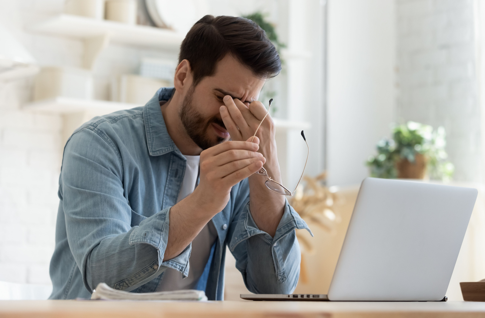 A young man sitting at a desk with his laptop and holding his glasses in his right hand as he rubs his eyes.