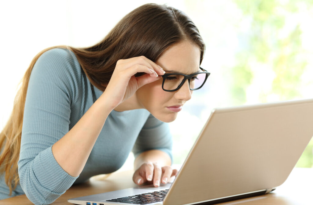 A woman holding her eyeglasses with her right hand, squinting to see clearly on her laptop